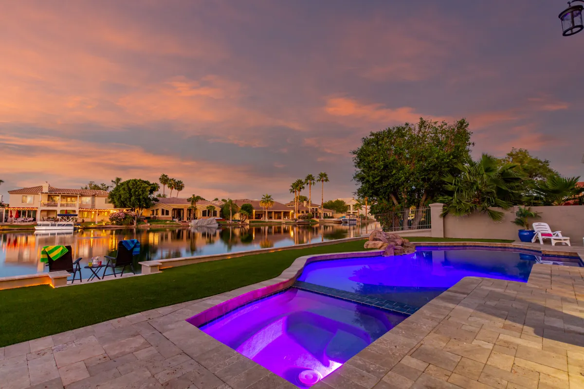 A glowing, heated pool stretches out in the foreground, flanked by vibrant, colorful chairs. Beyond the pool, tranquil lake waters reflect the sunset sky, creating a serene and luxurious atmosphere. The scene invites one to take a swim or unwind by the water while enjoying breathtaking waterfront views.