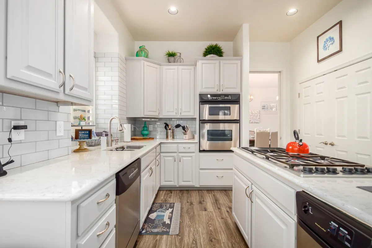 A contemporary kitchen featuring a spacious island at the center, surrounded by pristine white cabinetry and outfitted with stainless steel appliances. Decorative plants add a touch of greenery, and the overall space feels bright, modern, and functional. The layout emphasizes ample counter space and high-end fixtures, making it ideal for both serious cooking and casual gatherings.