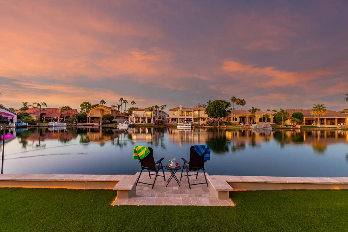 A serene waterfront view with two chairs and a small table for drinks, facing still water. Colorful towels drape over the chairs, and the reflections on the lake and warm hues suggest a tranquil sunset setting—ideal for relaxation and romantic moments.