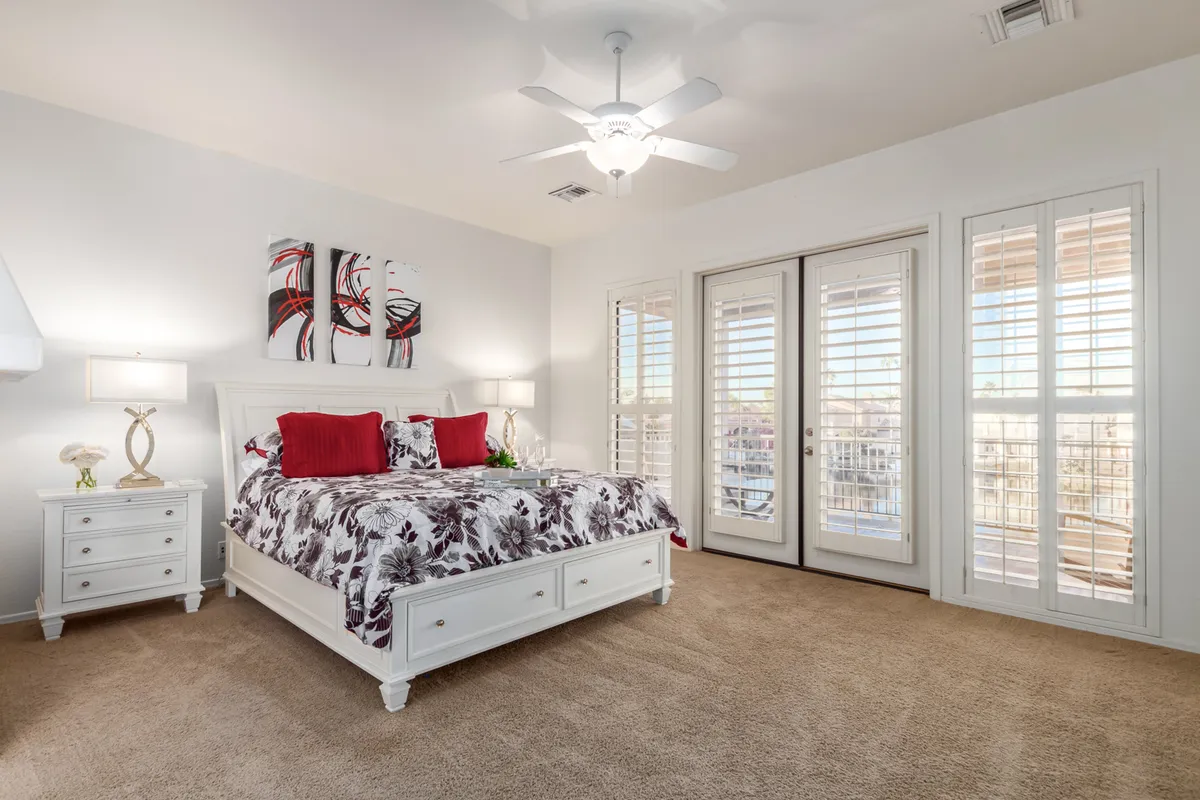 A beautifully appointed bedroom with floral bedding, abstract wall art, and nightstands. Double doors open to a private balcony, inviting sunlight and offering a glimpse of tranquil outdoor views.