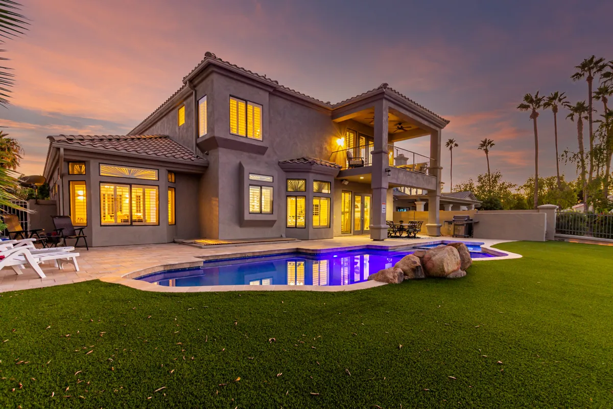 A luxurious backyard with a swimming pool, an upper balcony with elegant railings, and landscape lighting enhancing the space as evening approaches. The ambiance is both relaxed and upscale, with hints of outdoor dining and starlit gatherings.