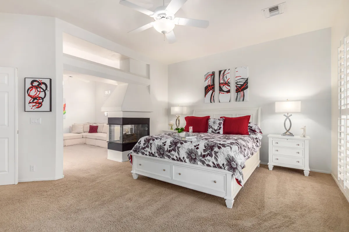 A serene master bedroom with a plush bed featuring floral bedding, a welcoming fireplace, tasteful artwork on the wall, and nightstands with warm lamps. Natural light pours in, creating a peaceful retreat that is both comfortable and elegant.