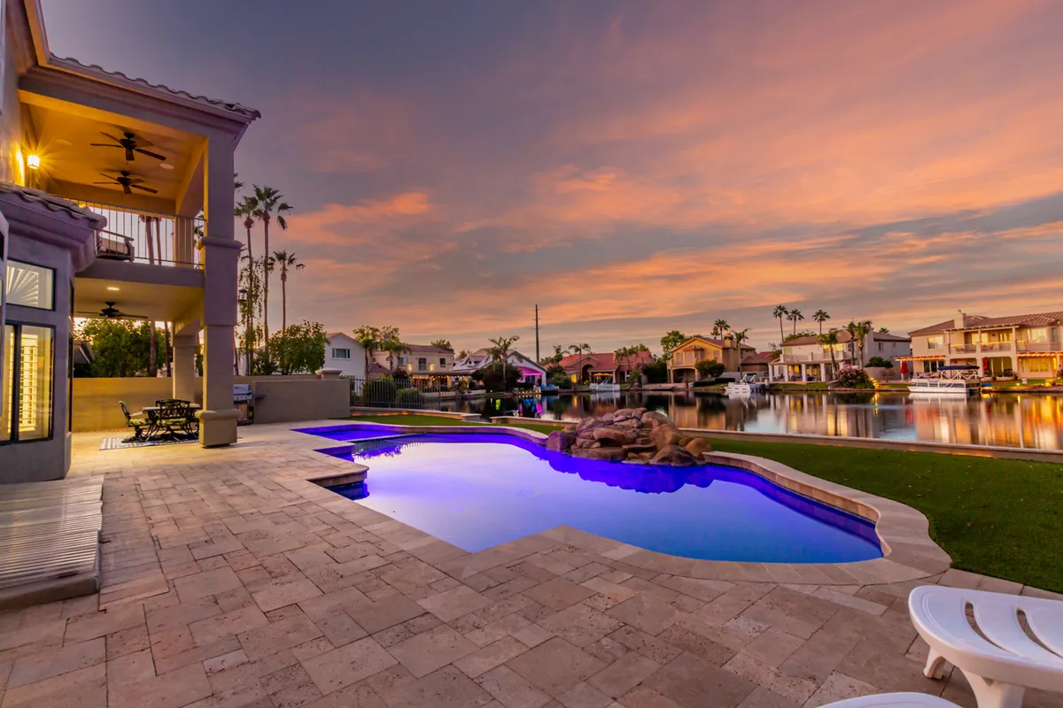 A stunning exterior view of a sparkling swimming pool with an integrated water feature, stylish patio seating, and a dramatic sunset sky. The serene, luxurious mood invites both relaxation and evening entertainment.