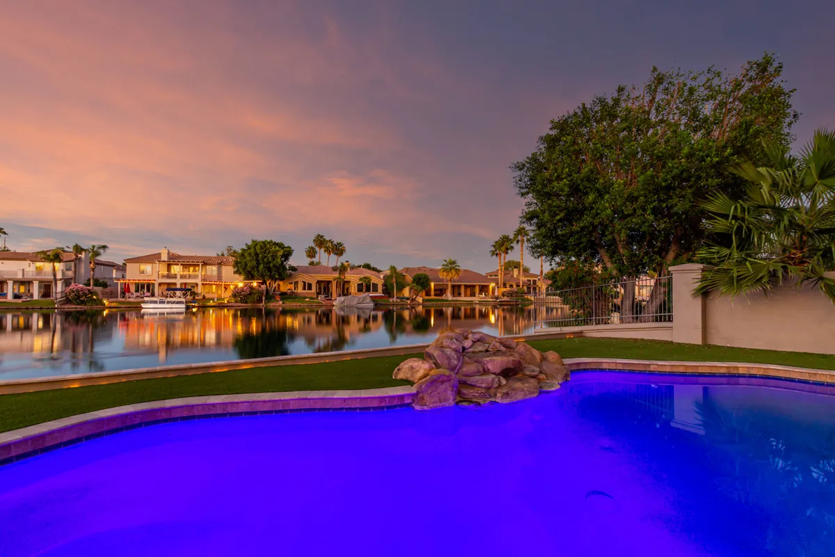 This image features a swimming pool illuminated with purple lighting, calm water, surrounding houses, and palm trees. The scene exudes a sense of community and relaxation, perfect for evening gatherings or family barbecues.