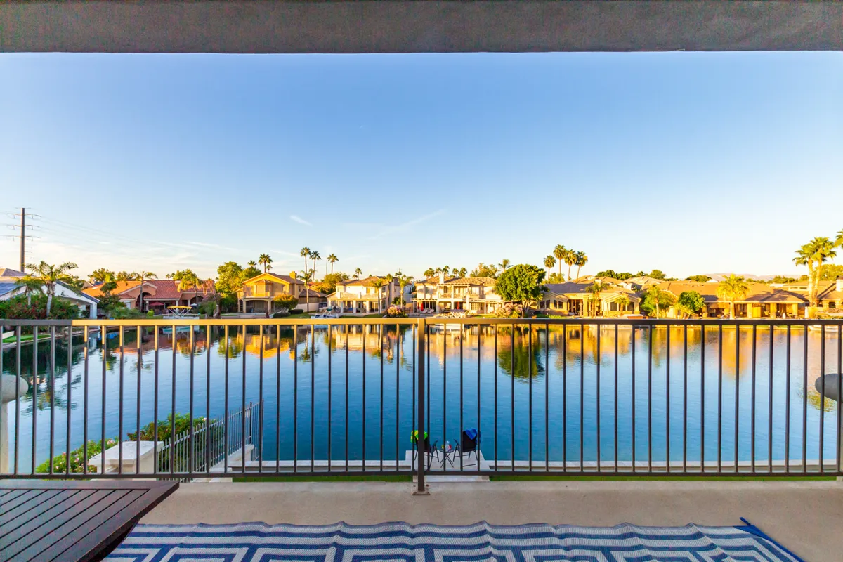 A scenic view with calm water in the foreground, colorful houses across the shore, and palm trees lining the waterfront. The tranquil mood suggests leisurely walks, intimate fishing moments, or group relaxation by the water.